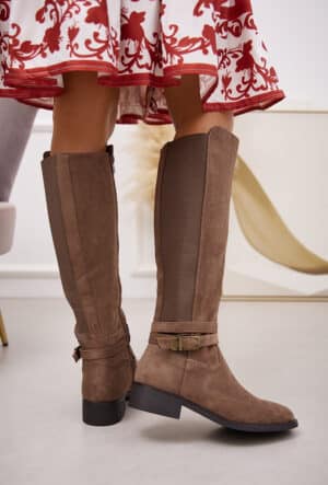 Avec une finition en suède taupe, ces bottes cavalières sont dotées d'une semelle discrète et de détails en métal au niveau de la cheville. L'ouverture zippée sur le côté permet de les enfiler facilement, et la bande élastique à l'arrière garantit un confort optimal au niveau du mollet.